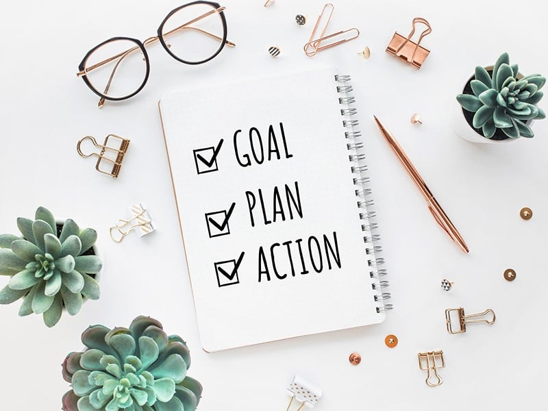 Desk top with a notepad surrounded by glasses, paperclips, plants and a pen. Written on the pad is a checklist saying Goal. Plan. Action.
